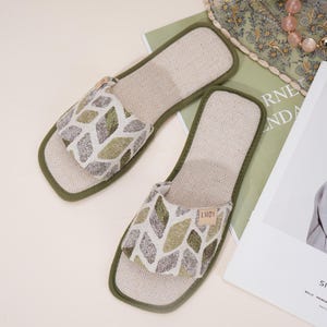 May include: A pair of open-toe slippers with a geometric pattern in shades of green, beige, and white. The slippers have a light beige interior and a dark green trim. The label "LMDY" is visible on one slipper.