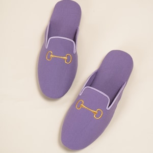 May include: A pair of lilac slippers featuring a gold-coloured bit design. The slippers have a rounded toe and a white trim around the opening. The slippers are made of a soft material.