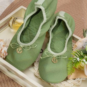 May include: A pair of olive green quilted fabric ballet slippers featuring a rounded toe, a soft elasticized collar, and a decorative thin cord bow at the front. Each slipper is accented with a small, circular gold-toned metal anchor charm.