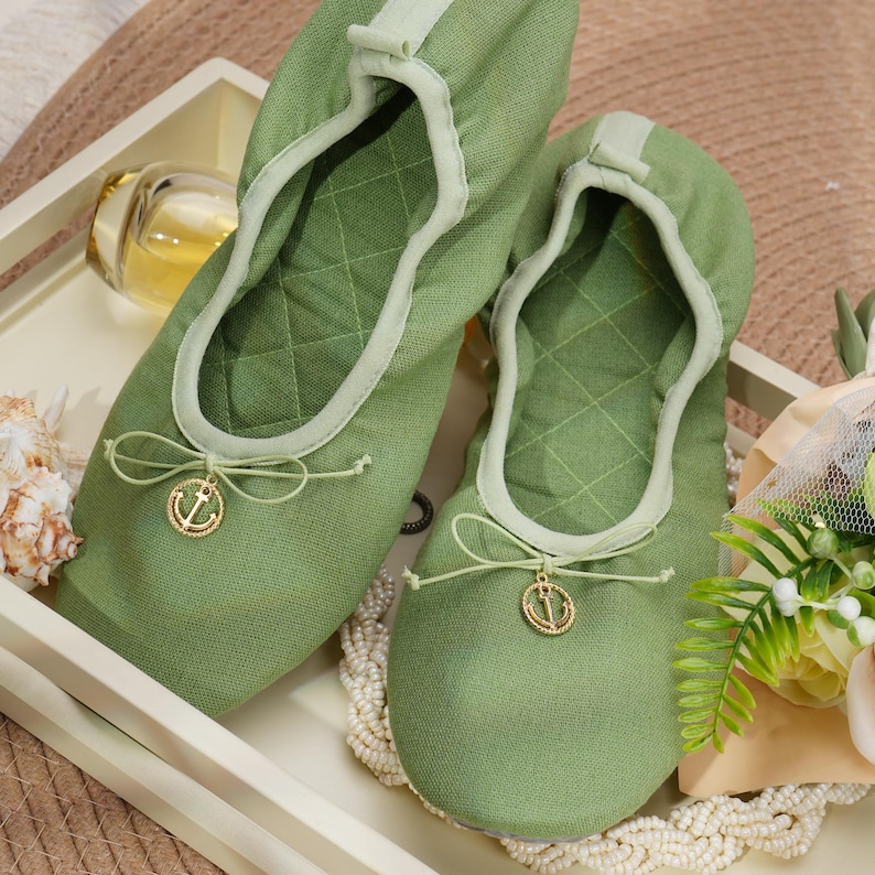 May include: A pair of olive green quilted fabric ballet slippers featuring a rounded toe, a soft elasticized collar, and a decorative thin cord bow at the front. Each slipper is accented with a small, circular gold-toned metal anchor charm.