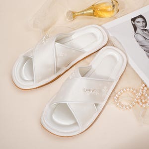 May include: White satin cross-strap slippers with floral embroidery and pearl accents. The slippers have a brown sole and are displayed with a perfume bottle and a fashion magazine. A string of pearls is also visible.