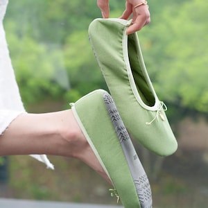 May include: A pair of sage green fabric ballet-style slippers featuring a rounded toe, a thin decorative drawstring bow at the front, and a grey textured rubber sole with a repeating geometric pattern for traction on the bottom of the footwear.