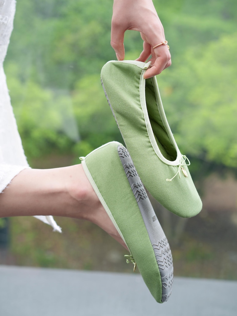 May include: A pair of sage green fabric ballet-style slippers featuring a rounded toe, a thin decorative drawstring bow at the front, and a grey textured rubber sole with a repeating geometric pattern for traction on the bottom of the footwear.