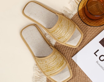 Può includere: Un paio di pantofole intrecciate beige e marroni con interno colorato chiaro. Le pantofole sono posizionate su un tappetino intrecciato con una tazza di tè e una rivista sullo sfondo. Le pantofole hanno un design a punta quadrata.