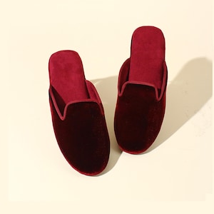 May include: A pair of burgundy velvet slippers with a closed toe and a back that is open. The slippers have a soft, plush feel.