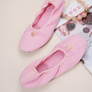 May include: A pair of light pink ballet flats featuring a rounded toe, a gathered elastic topline, and a small gold-toned anchor charm attached to a thin decorative bow at the center of the vamp. The shoes have a soft, fabric-like exterior.