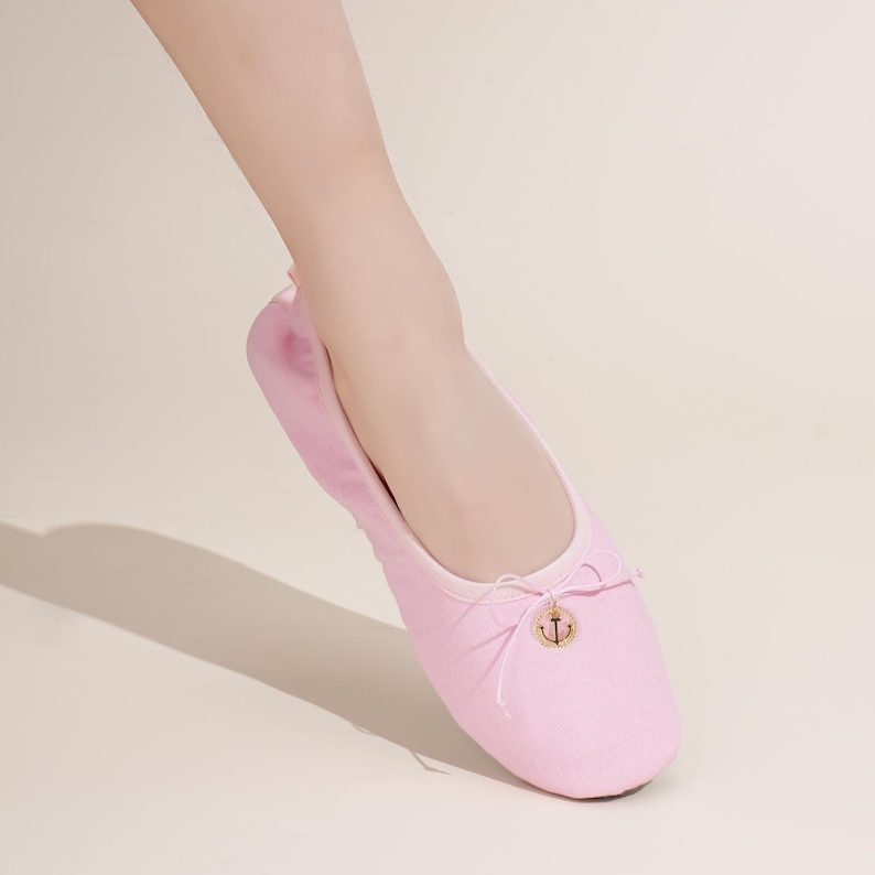 May include: A light pink ballet flat shoe featuring a rounded toe and a thin drawstring tie at the front. A small, circular gold-toned charm with an anchor emblem is attached to the center of the bow, adding a metallic detail to the fabric.