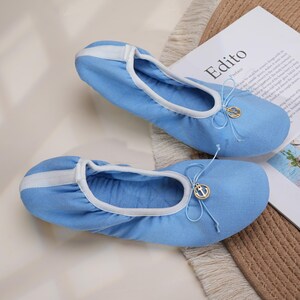 May include: A pair of light blue fabric ballet flats featuring a white trim along the collar and a white vertical stripe on the heel. Each shoe includes a decorative light blue bow at the front adorned with a small gold-toned circular anchor charm.