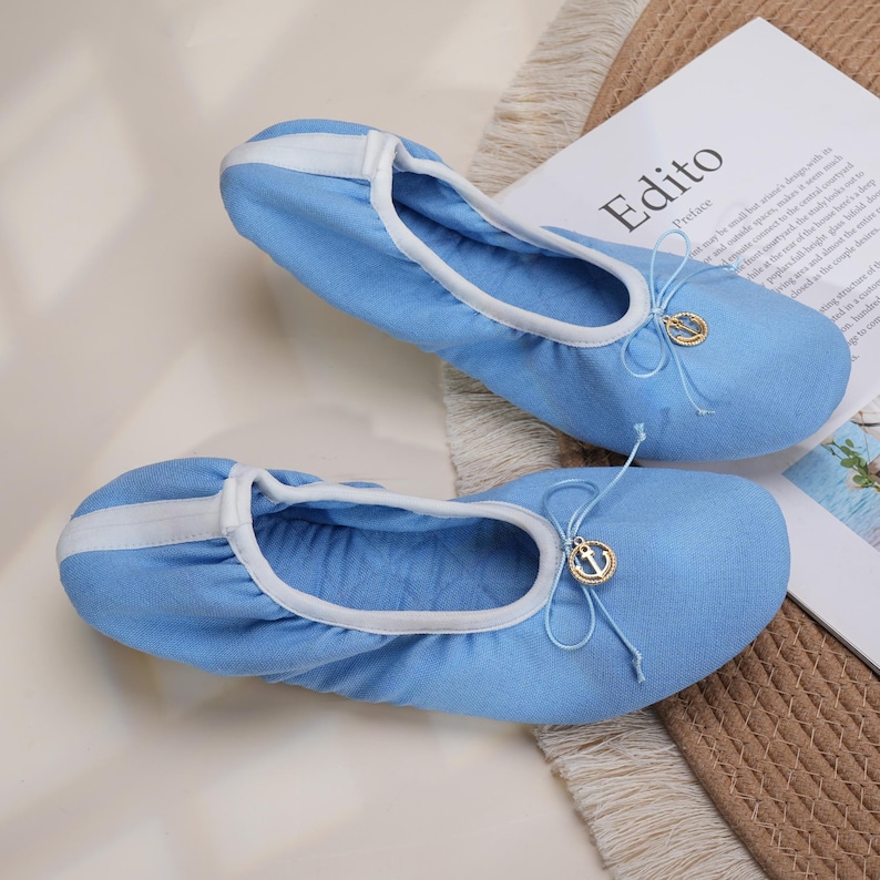 May include: A pair of light blue fabric ballet flats featuring a white trim along the collar and a white vertical stripe on the heel. Each shoe includes a decorative light blue bow at the front adorned with a small gold-toned circular anchor charm.