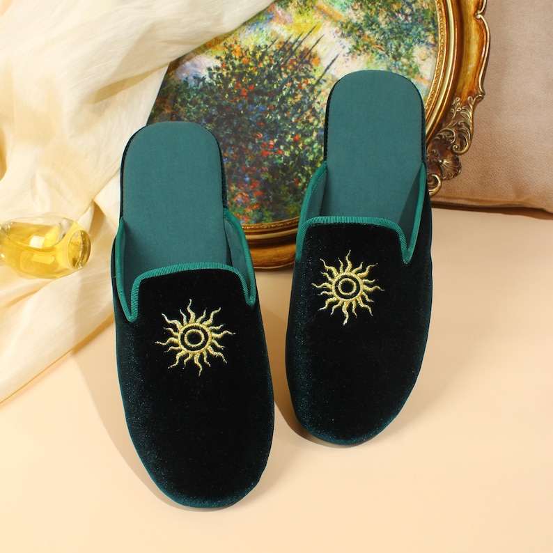 May include: A pair of dark green velvet slippers featuring a gold embroidered sun design on the top of each shoe. The slippers have a matching dark green interior lining and a teal green trim along the opening of the slip-on footwear design.