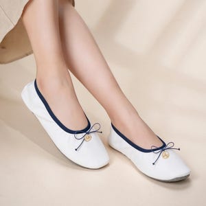 May include: A pair of white ballet flats featuring a dark blue trim along the collar and a small decorative bow at the front. Each shoe is accented with a circular gold-toned charm attached to the center of the bow, providing a subtle detail.