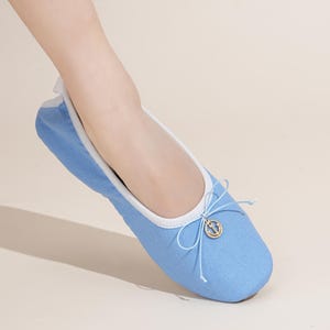 May include: A light blue ballet flat shoe featuring a white trim along the collar and a delicate light blue bow at the front. A small gold-toned circular charm with an anchor emblem is attached to the center of the bow on the upper shoe.