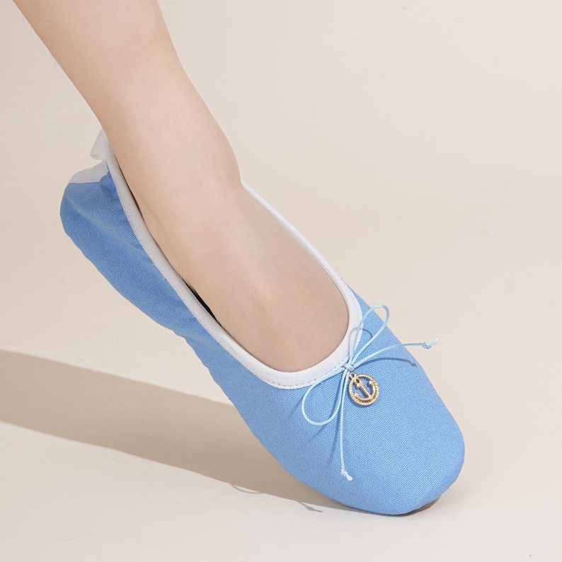 May include: A light blue ballet flat shoe featuring a white trim along the collar and a delicate light blue bow at the front. A small gold-toned circular charm with an anchor emblem is attached to the center of the bow on the upper shoe.