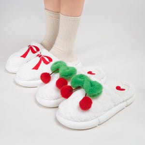 Pu&ograve; includere: Pantofole in peluche bianche con ricami a fiocco rosso e un paio con accenti di ciliegia. Le pantofole hanno una suola spessa e sono esposte con calzini beige.