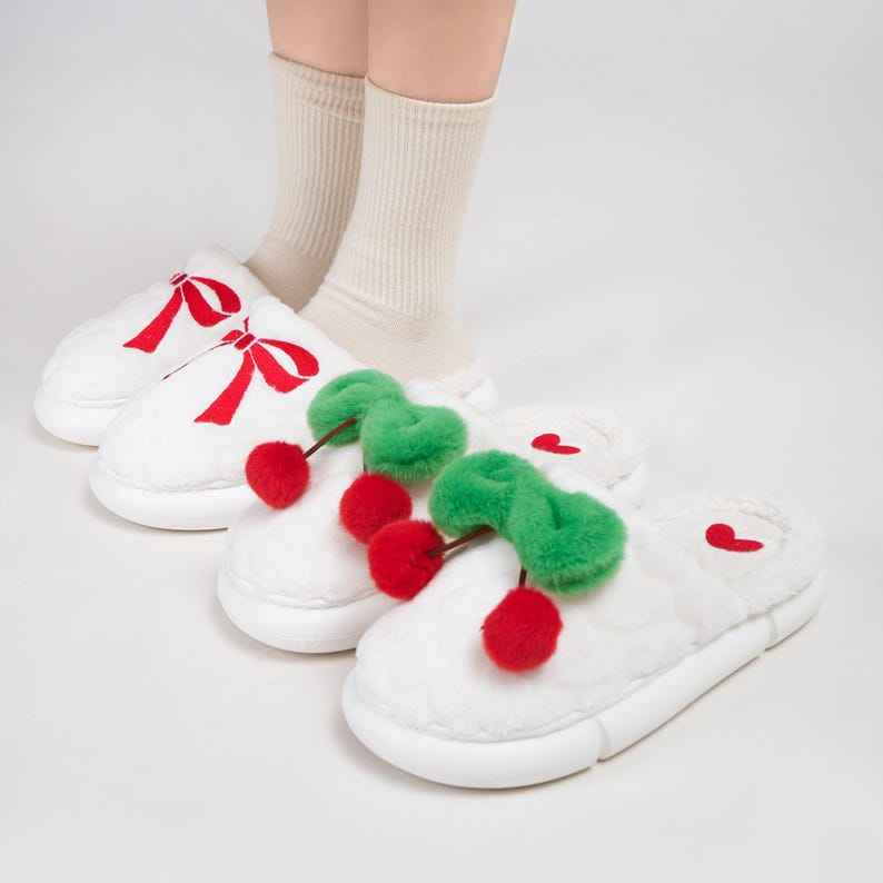 Pu&ograve; includere: Pantofole in peluche bianche con ricami a fiocco rosso e un paio con accenti di ciliegia. Le pantofole hanno una suola spessa e sono esposte con calzini beige.
