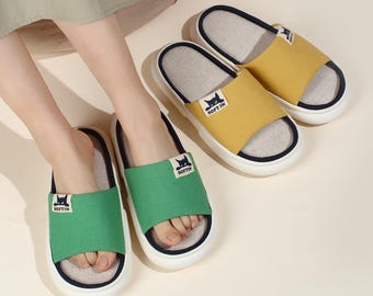 May include: A pair of green and a pair of yellow open-toe slippers. Each slipper has a white sole with a navy blue trim, and a fabric strap with a cat logo and the text "SOFT IN". The slippers are designed for indoor use.
