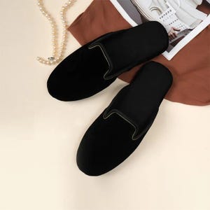 Puede incluir: Un par de zapatos negros de terciopelo sin cordones con ribete dorado. Los zapatos tienen forma ovalada y están sobre una superficie de color claro con una tela marrón y un collar de perlas en el fondo.