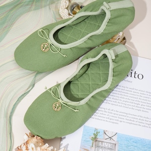 May include: A pair of sage green ballet flats featuring a quilted interior lining, a rounded toe, and a decorative gold-toned anchor charm attached to a thin drawstring bow on the front of each shoe. The shoes have a soft, flexible construction.