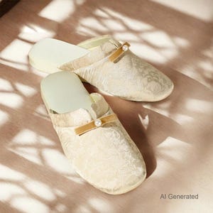 K&ouml;nnte beinhalten: Ein Paar cremefarbene Slipper mit einem dezenten floralen Jacquardmuster. Jeder Slipper ist mit einem horizontalen beigen Samtriemen &uuml;ber dem Vorderblatt verziert, der durch eine einzelne, mittig platzierte wei&szlig;e Perle akzentuiert wird.
