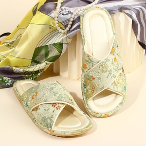 May include: A pair of light green floral patterned slippers with a cream-coloured interior. The slippers have a cross-strap design and are set against a backdrop of a silk scarf and a pearl necklace. The slippers are a comfortable footwear option.