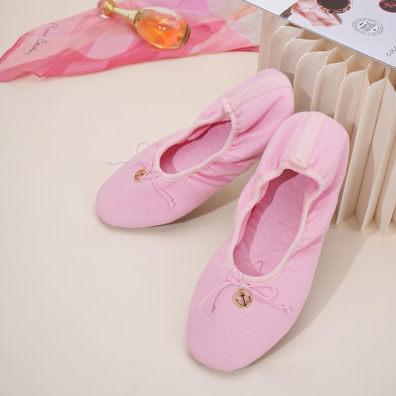 May include: A pair of light pink fabric ballet slippers featuring a rounded toe, an elasticized topline, and a decorative thin drawstring bow at the front. Each slipper is accented with a small, circular gold-toned charm featuring an anchor design.