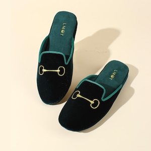 May include: A pair of dark green velvet slippers featuring a gold-coloured horsebit detail on the top. The slippers have a matching green trim and the text LMOY is embroidered in gold on the inner sole of each slipper, which are slip-on style.