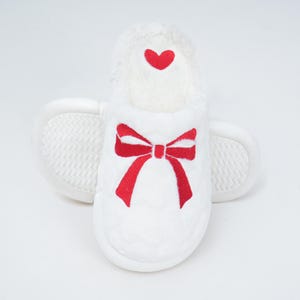Pu&ograve; includere: Pantofola in peluche bianca con un design a fiocco e cuore rosso. La pantofola ha un interno morbido e strutturato e una suola antiscivolo. Il fiocco &egrave; ricamato sul davanti e un piccolo cuore &egrave; ricamato in alto.