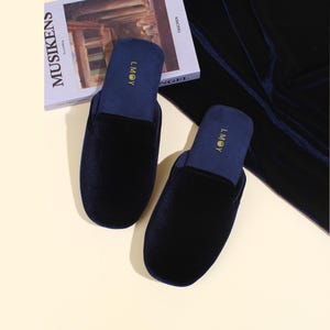May include: A pair of dark navy blue velvet slippers with a rounded toe and a slip-on design. The interior footbed is a lighter shade of blue and features the gold embroidered text LMOY. The slippers have a soft, plush texture throughout.