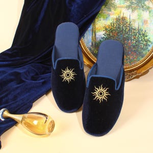 May include: A pair of dark navy blue velvet slippers featuring a gold embroidered sun design on the top. The slippers have a matching navy blue interior lining and trim, with a rounded toe silhouette and a slip-on style construction.