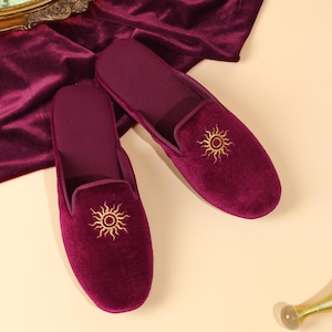 May include: A pair of deep burgundy velvet slippers featuring a gold embroidered sun motif on the top of each shoe. The slippers have a closed-toe design with a smooth, soft texture and a matching burgundy velvet interior lining.