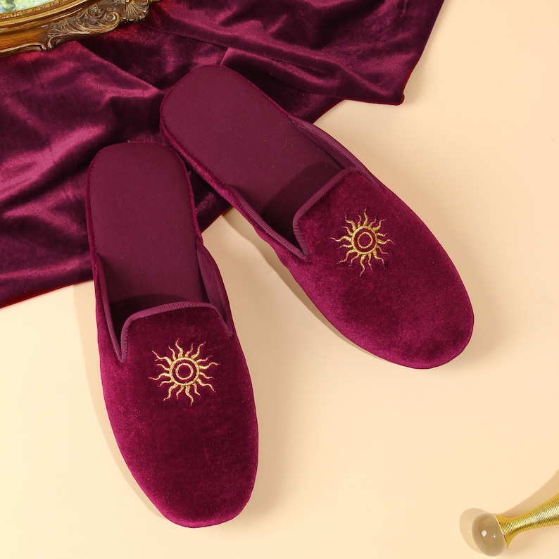 May include: A pair of deep burgundy velvet slippers featuring a gold embroidered sun motif on the top of each shoe. The slippers have a closed-toe design with a smooth, soft texture and a matching burgundy velvet interior lining.