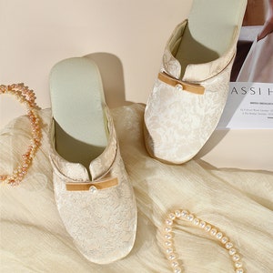 K&ouml;nnte beinhalten: Ein Paar cremefarbene Slipper mit einem tonalen floralen Jacquardmuster, einer kleinen beigen Samtschleife auf dem Vorderblatt und einem einzelnen wei&szlig;en Kunstperlenakzent. Die Slipper haben ein hellgr&uuml;nes Innenfutter und eine runde Zehenpartie.