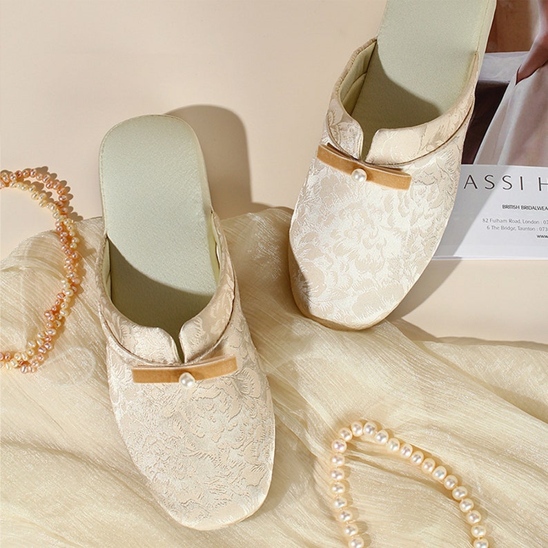 K&ouml;nnte beinhalten: Ein Paar cremefarbene Slipper mit einem tonalen floralen Jacquardmuster, einer kleinen beigen Samtschleife auf dem Vorderblatt und einem einzelnen wei&szlig;en Kunstperlenakzent. Die Slipper haben ein hellgr&uuml;nes Innenfutter und eine runde Zehenpartie.
