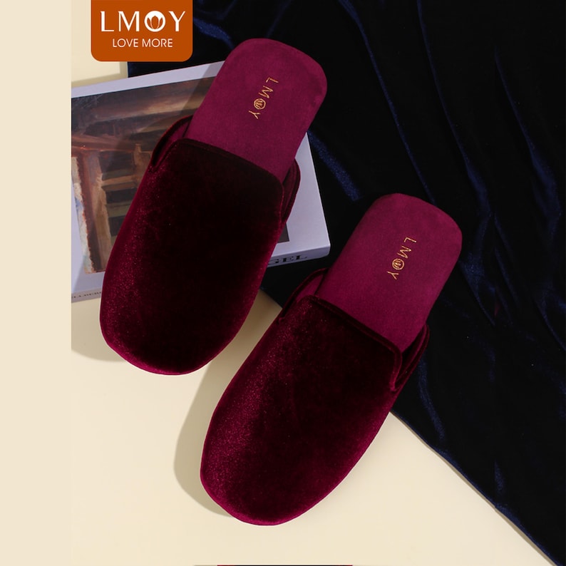 May include: A pair of deep burgundy velvet slippers with a rounded toe design. The interior footbed features the gold text LMOY printed near the heel. The slippers have a soft, plush texture and a solid dark red colour throughout the exterior surface.