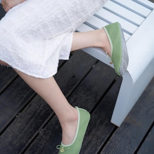 May include: A pair of sage green ballet flats featuring a rounded toe, a light green trim, and a small decorative bow at the front with a gold-toned circular charm. The soles are grey with a textured, non-slip geometric pattern for added grip.