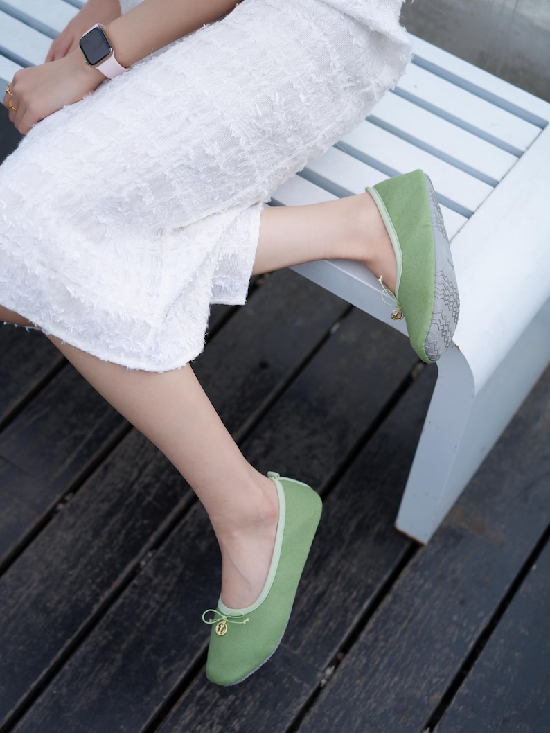 May include: A pair of sage green ballet flats featuring a rounded toe, a light green trim, and a small decorative bow at the front with a gold-toned circular charm. The soles are grey with a textured, non-slip geometric pattern for added grip.