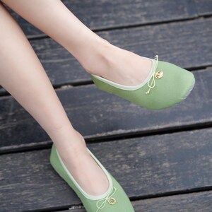 May include: A pair of sage green ballet flats featuring a rounded toe, a light green trim along the collar, and a delicate drawstring bow detail at the front. Each shoe is accented with a small, circular gold-toned charm attached to the bow.