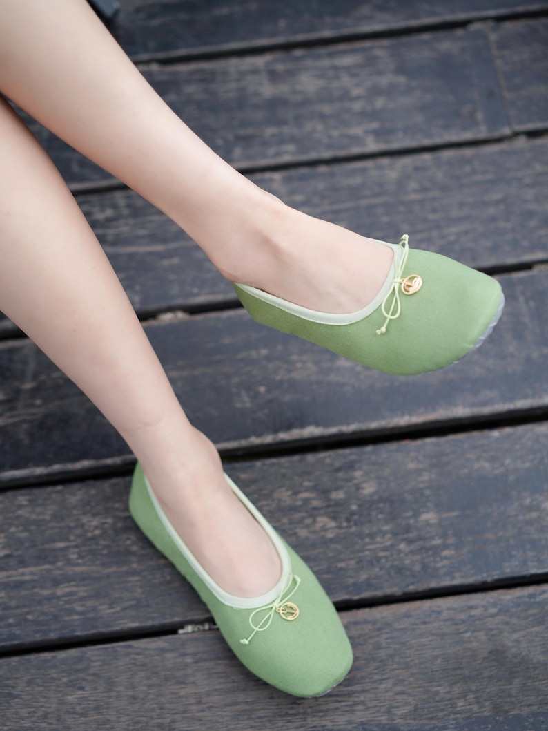 May include: A pair of sage green ballet flats featuring a rounded toe, a light green trim along the collar, and a delicate drawstring bow detail at the front. Each shoe is accented with a small, circular gold-toned charm attached to the bow.