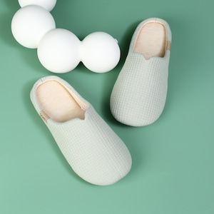 May include: A pair of light green knitted slippers with a soft, beige lining. The slippers have a simple, slip-on design.