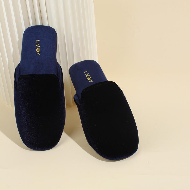 May include: A pair of dark navy blue velvet slippers with a rounded toe and a slip-on design. The interior footbed features the gold text LMOY printed near the heel area. The slippers have a smooth, soft texture and a solid dark blue colour.