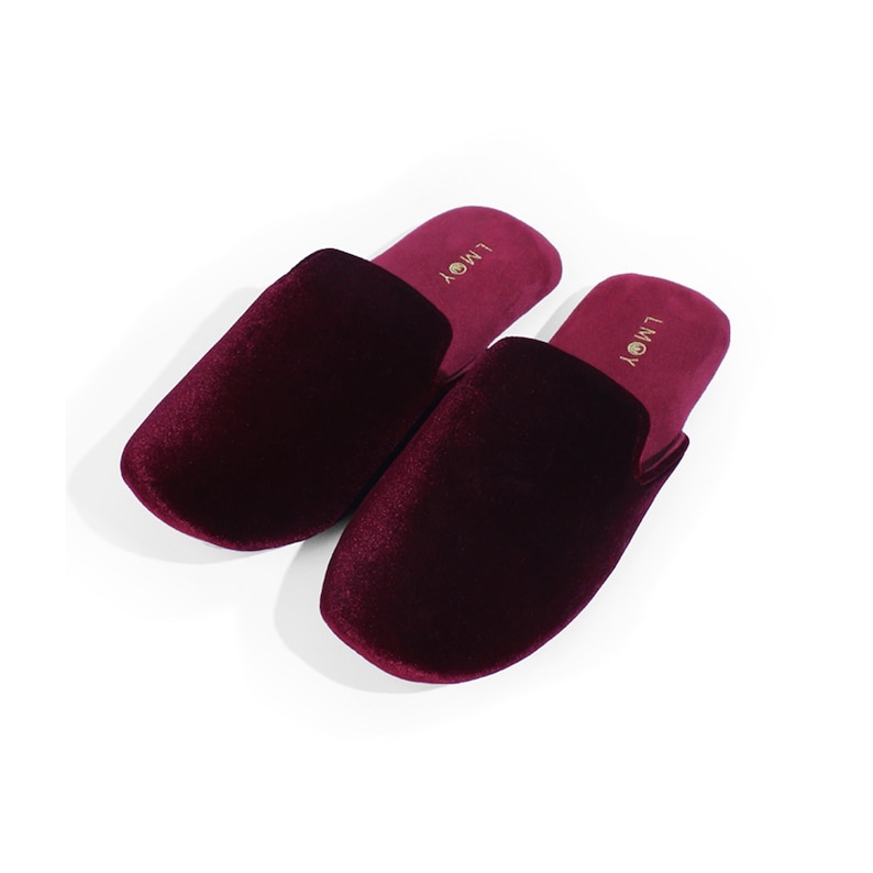 May include: A pair of deep burgundy velvet slippers with a closed-toe design. The interior footbed is lined in matching burgundy fabric and features the gold-coloured text LMOY printed near the heel area of each slipper.
