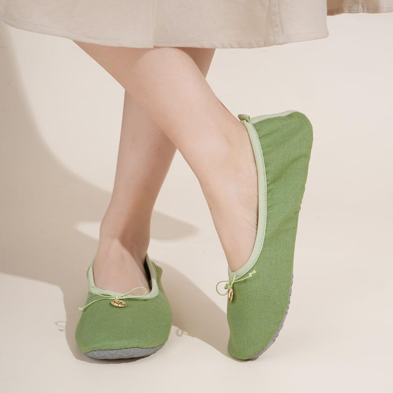 May include: A pair of sage green fabric ballet-style slippers featuring a rounded toe, a thin drawstring tie at the front with a small gold-toned circular charm, a light green trim along the collar, and a textured grey rubber sole.