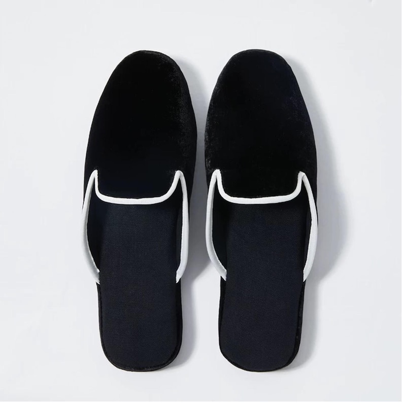 May include: A pair of black velvet slippers with white piping around the edges. The slippers have a closed toe and a slip-on design.