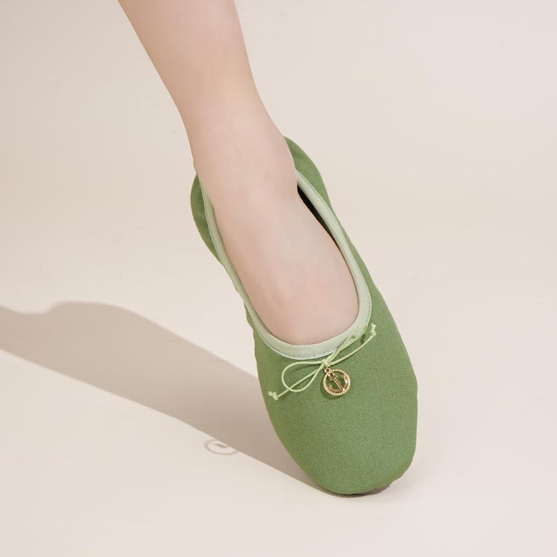 May include: A sage green canvas ballet flat shoe featuring a light green trim around the collar and a delicate drawstring bow at the front. A small, circular gold-toned metal charm with a detailed emblem hangs from the center of the bow.