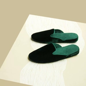 May include: A pair of dark green velvet slippers with a closed toe and an open back. The slippers are on a white surface with a wavy pattern.