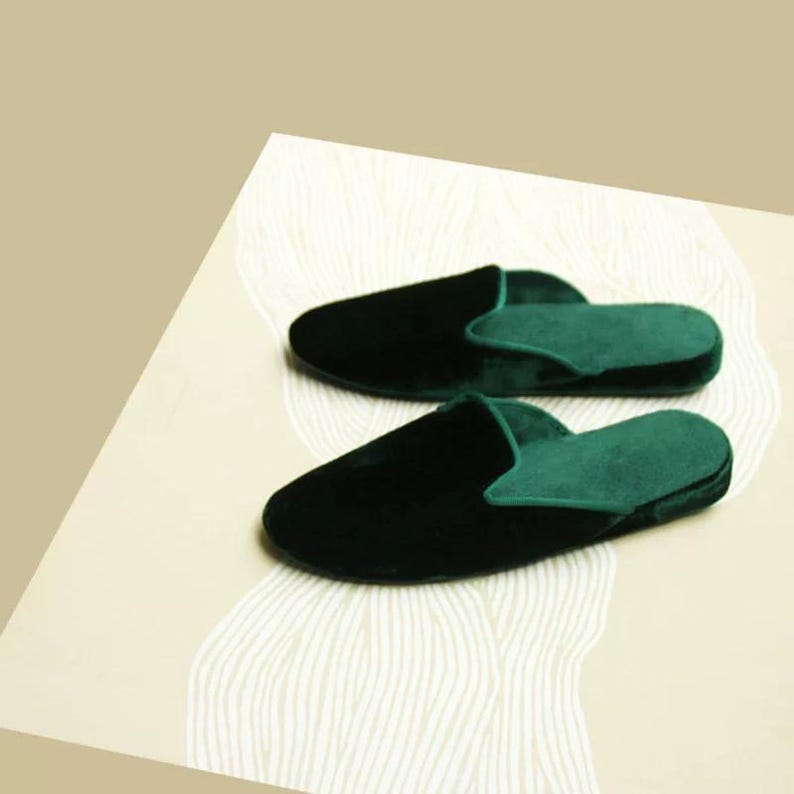 May include: A pair of dark green velvet slippers with a closed toe and an open back. The slippers are on a white surface with a wavy pattern.