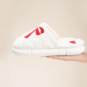 Pu&ograve; includere: Pantofola bianca e soffice con un design a nastro rosso sulla parte superiore. La pantofola ha una suola bianca e un piccolo cuore rosso sul tallone. Progettata per l'uso in casa.