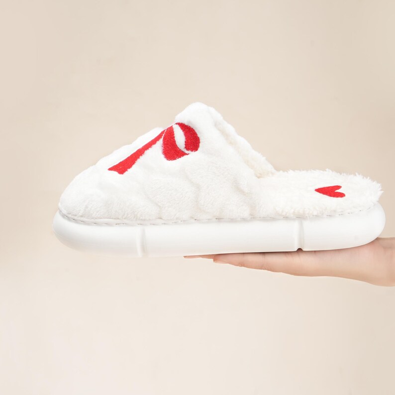 Pu&ograve; includere: Pantofola bianca e soffice con un design a nastro rosso sulla parte superiore. La pantofola ha una suola bianca e un piccolo cuore rosso sul tallone. Progettata per l'uso in casa.