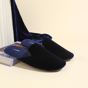 May include: A pair of deep navy blue velvet slippers with a closed-toe design. The interior footbed features the gold text LMOY printed on the heel. The slippers are crafted from a soft, plush fabric with a smooth texture and a rounded toe shape.