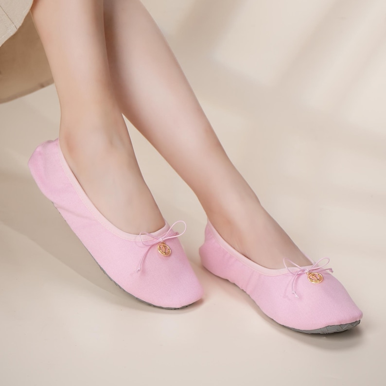 May include: A pair of light pink ballet-style slippers featuring a soft fabric upper, a rounded toe, and a delicate decorative bow at the center of the vamp. Each slipper is accented with a small, circular gold-toned charm attached to the bow.